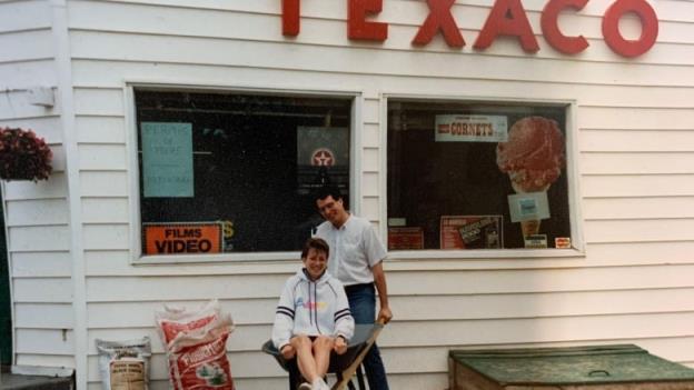 A couple smiles at the camera standing in front of a white building. A woman sits in a wheel barrow and a man is behind her. 