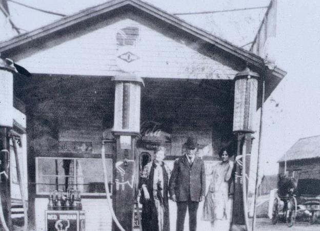 A black and white photo of a man and two women standing in front of a gas pump and a building. 