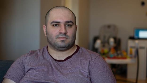 A man sits in a small apartment with children's toys behind him.