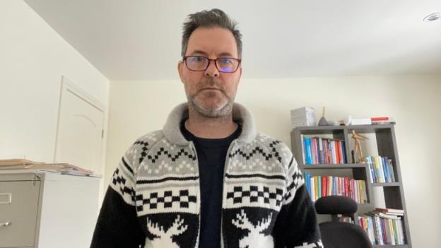 A man wearing a moose sweater and glasses stands in his home office.