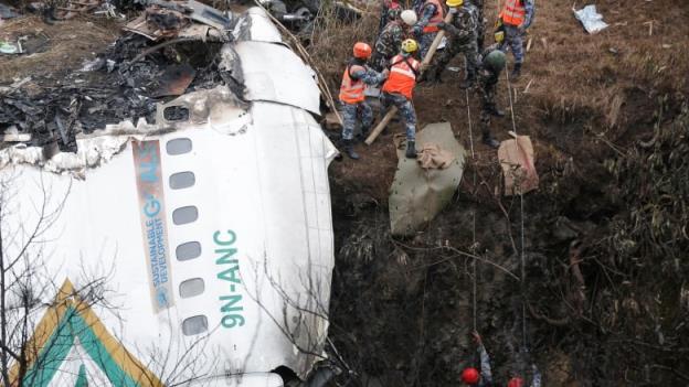 Part of a passenger plane fuselage is seen resting on a cliff. Rescuer workers, using ropes, attempt to reach the wreckage. 