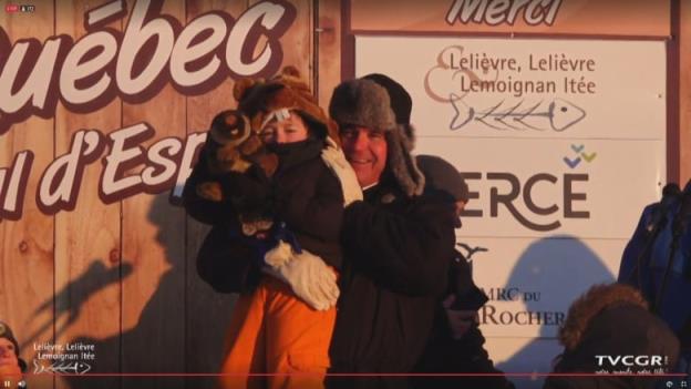 A man hoisting up a child wearing a groundhog hat. 