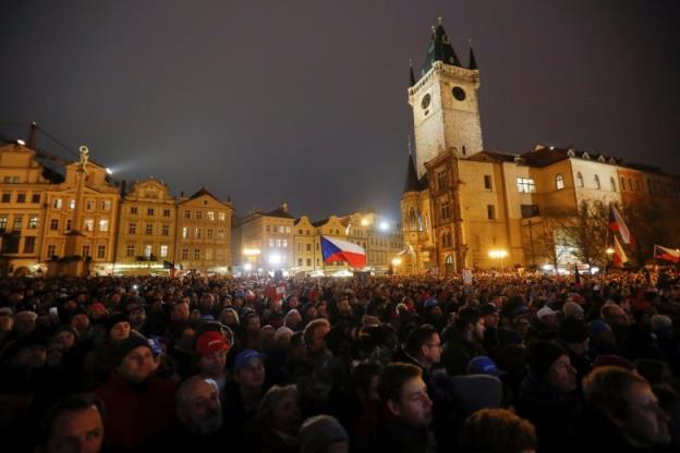 A massive crowd gathers in front of brightly lit buildings, a lone flag rising from the sea of people.