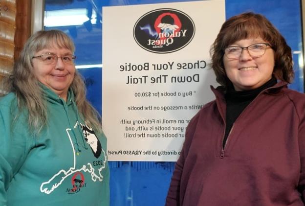 Two women stand in front of a Yukon Quest sign.