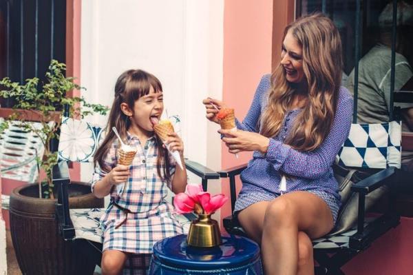 Mom with her 6 years old daughter walking along city street and eating ice cream in front of the outdoor cafe. Good relations of parent and child. Happy moments together.