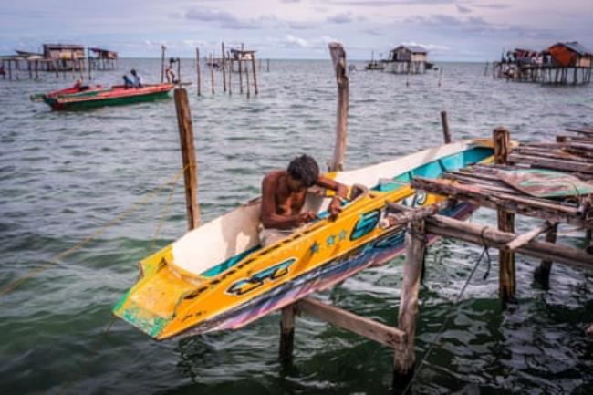 A man working on a simple boat, sitting on the stilts of his house.