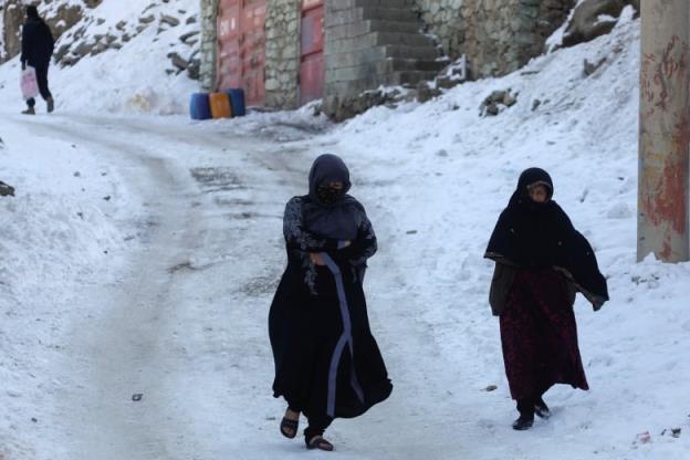 Two women wearing head scarves and long clothes walk down a snowy street in Kabul, Afghanistan.