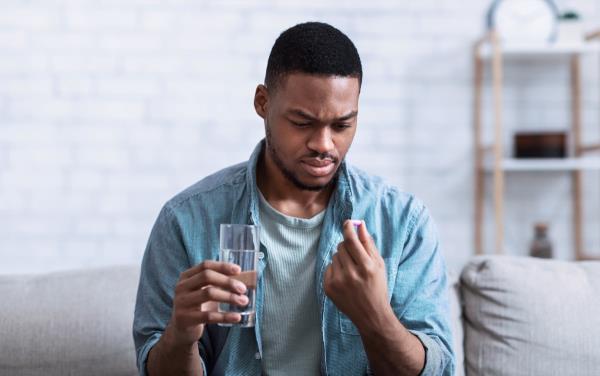 Man Holding Pill And Glass Of Water Taking Medicine Sitting On Sofa At Home.