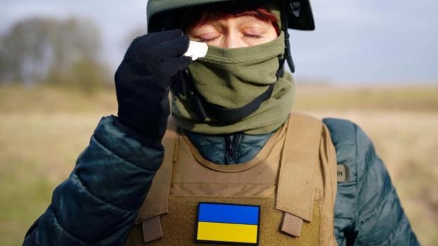 A soldier in full combat gear including a helmet holds up a tissue to her eyes, which are closed. A Ukrainian flag is on the middle of the front of her clothing.