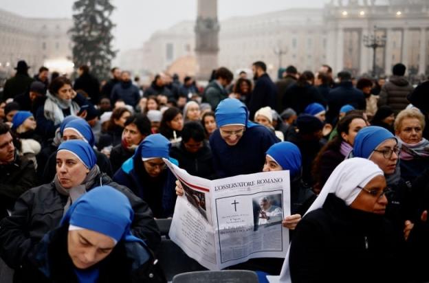 Nuns in blue veils gather in a square at dawn. Two of them read from a newspaper with bearing the photo of pope emeritus Benedict XVI.